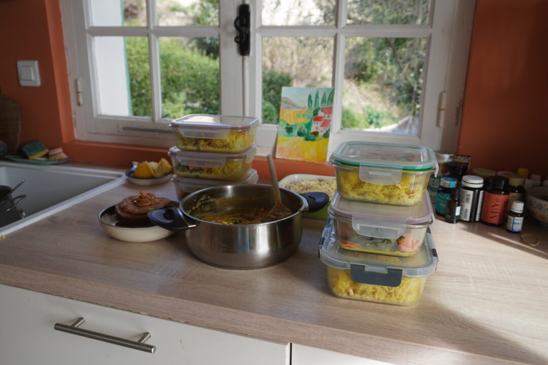 Vue d'ensemble d'un kit de batch cooking complet sur table en bois : plusieurs contenants en verre empilés, un plat ouvert et des tomates cerises fraîches.